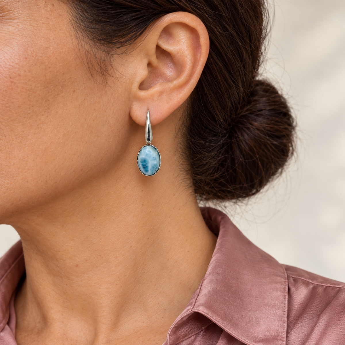 Model with chestnut hair in a low bun and dusty rose blouse wearing oval Larimar earrings; approximate scale mockup.