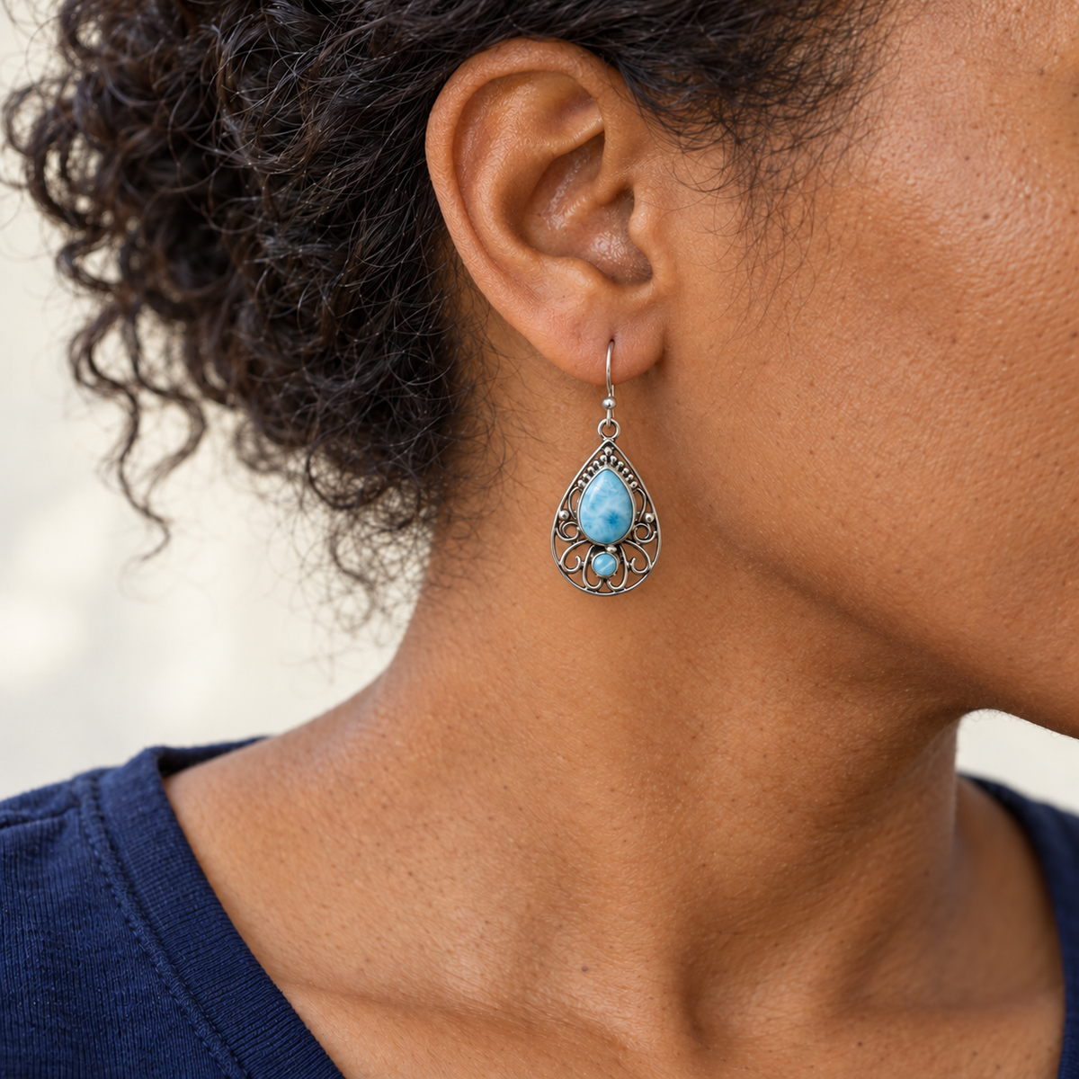 Model with pulled-back curls and navy shirt wearing a teardrop Larimar earring with silver scrollwork; approximate scale mockup.
